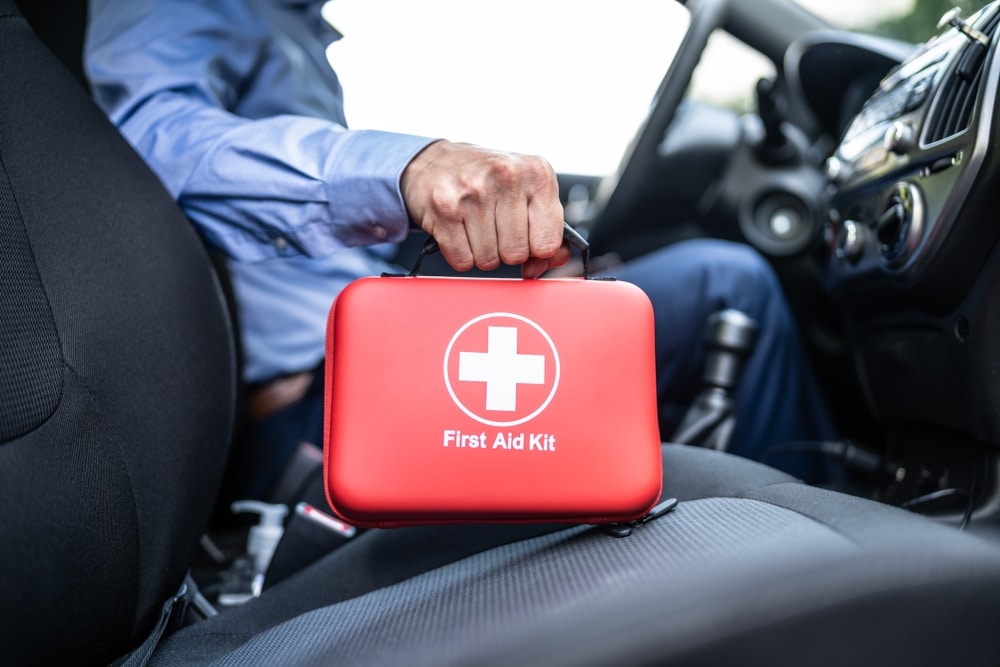 A person in business attire sitting in a car holds a red first aid kit with a white cross on it, placed on the front passenger seat.